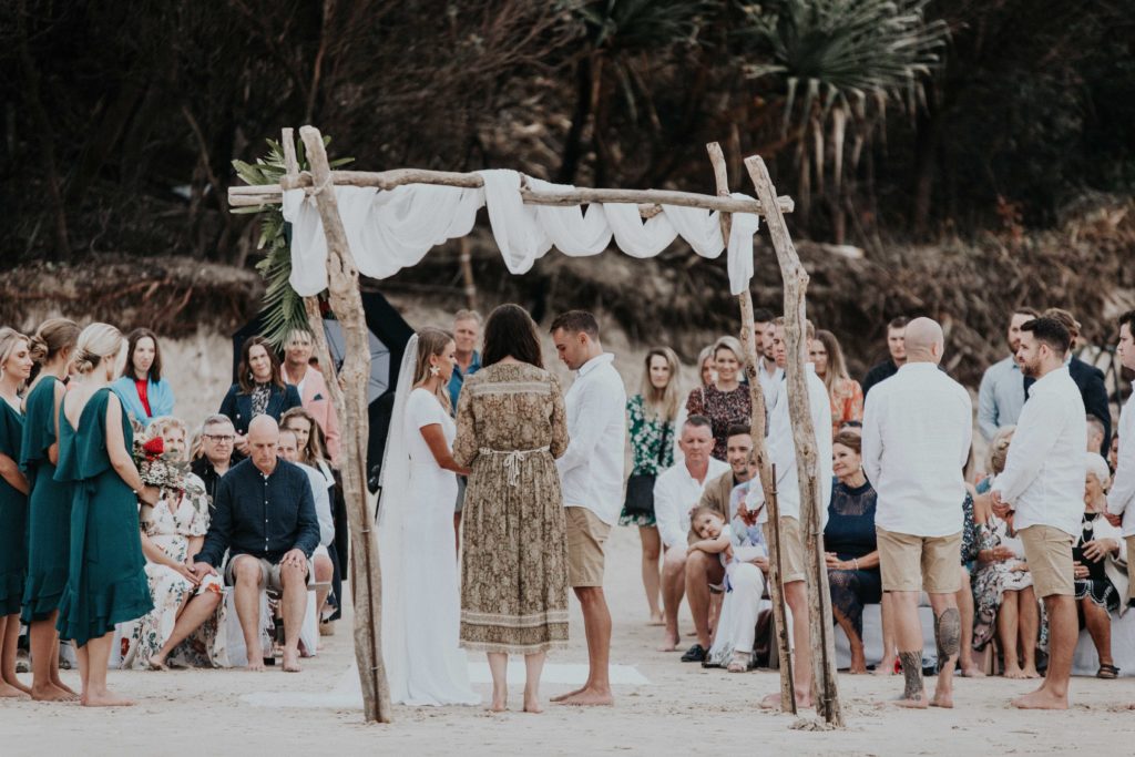 Barefoot and grounded - Jasmine and Ben holding hands during their Byron Bay beach wedding ceremony. All of the guests had their bare feet in the sand.