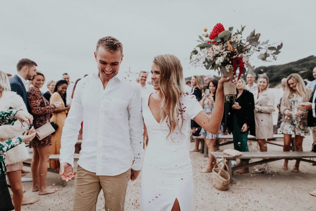 Jasmine and Ben holding hands recessing back down the aisle post-ceremony. The guests throwing eco-confetti for their beach wedding ceremony.
